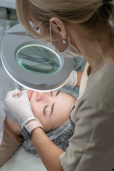 Cosmetologist performing a facial treatment. Close-up shows hands working on a client's face under a magnifying lamp. Precise procedure in progress.