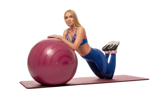 Side view of fit female athlete in sportswear sitting on fitness ball and looking at camera against white background