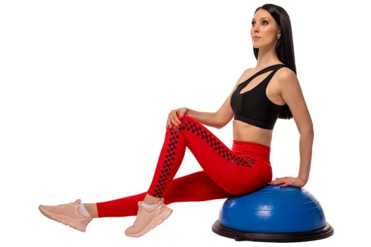 Full body side view of slim young female in sportswear and sneakers sitting on blue fitness ball and looking at camera isolated on white background