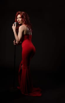 young graceful female vocalist in elegant red maxi dress with long curly ginger hair singing song in retro microphone and looking at camera