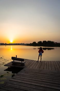 A scenic lakeside view at sunset with a woman standing on a wooden pier, featuring a wooden bench and golden reflections on calm water.