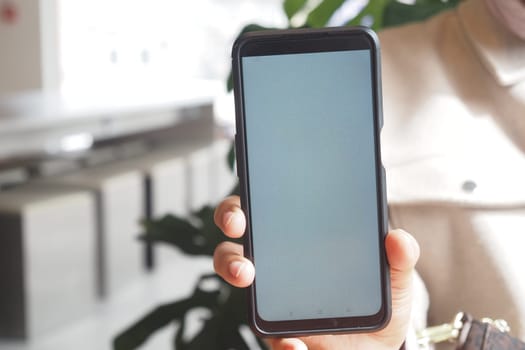 A person shows a blank smartphone screen while indoors surrounded by greenery.