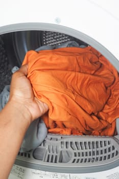 A hand places a vibrant orange shirt into a washing machine for cleaning.