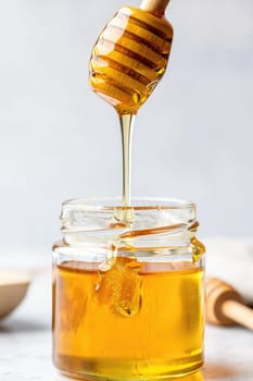A wooden dipper is immersed in a glass jar filled with golden honey. The honey drips slowly from the dipper, creating a mesmerizing flow. Soft lighting illuminates the bright kitchen.