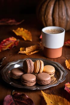 Assorted macarons sit on a silver platter next to a warm drink in a cozy setting filled with autumn leaves. The earthy colors evoke a festive, inviting atmosphere perfect for fall.