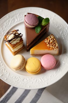 A variety of tempting desserts are arranged on a white plate, showcasing cakes and macarons. The setting is cozy with natural light highlighting the sweet treats.