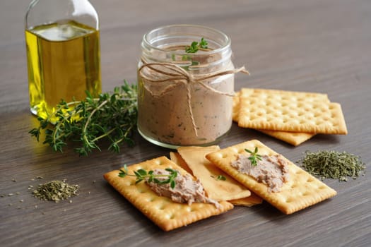 Savory spread made from blended ingredients sits in a jar next to crackers and fresh herbs. Olive oil is also present, completing the rustic presentation on a wooden table.