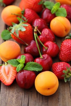 An assortment of colorful summer fruits including cherries, strawberries, apricots, and mint leaves is neatly spread on a rustic wooden surface. The fruits display vivid colors and textures.