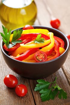 Brightly colored strips of red and yellow bell peppers are mixed with cherry tomatoes and fresh parsley in a bowl. Olive oil and rustic wood create a warm atmosphere.