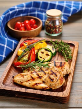 A delicious grilled chicken breast is plated alongside colorful grilled vegetables including bell peppers and zucchini. Fresh herbs enhance the dish in a warm and inviting kitchen environment.