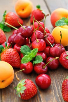 A vibrant collection of fresh cherries, strawberries, and apricots arranged on a rustic wooden table. Green mint leaves add a touch of color to the display. This is a summer harvest bounty.