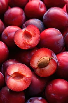 Bright red plums are clustered closely together, showcasing their shiny skin. Some are sliced in half, exposing their juicy interior with a central pit. A display of freshness inviting tasting.