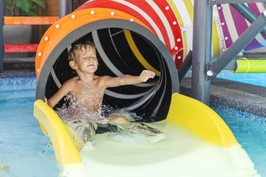 Wet boy exiting colorful tube water slide into shallow pool, water splashing around his legs and arms as he holds one arm up and closes his eyes