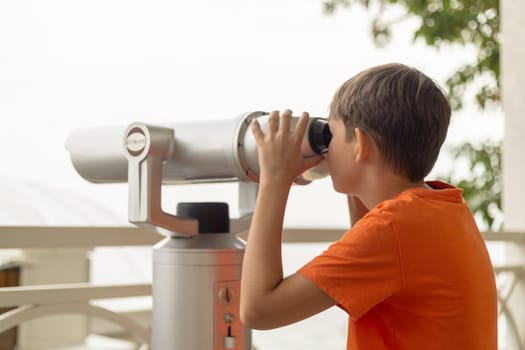Boy in orange shirt looking through a mounted public viewing telescope on a terrace with railing and distant view obscured by fog or haze