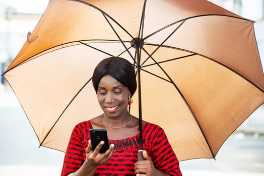 mature woman in traditional clothes standing looking at mobile phone smiling with umbrella over her head.