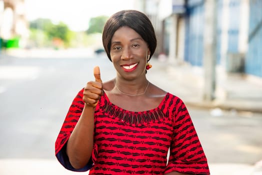 mature woman in traditional outfit standing outdoors looking at camera smiling and making hand gesture.