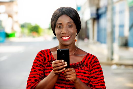 happy african adult business woman, dressed in red loincloth, walking outdoors in the street, using mobile phone