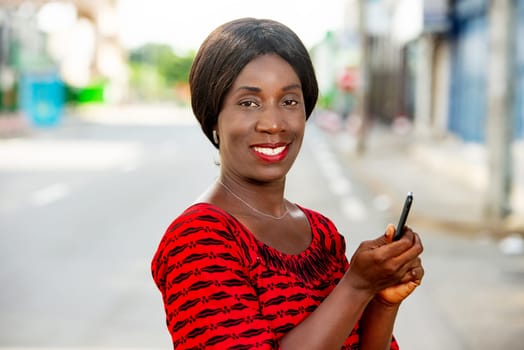 happy african adult business woman, dressed in red loincloth, walking outdoors in the street, using mobile phone