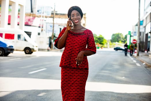 Happy african adult business woman, dressed in red loincloth, is communicating on the phone while walking in the street