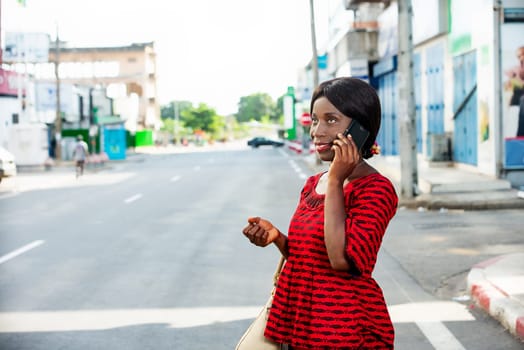 Happy african adult business woman, dressed in red loincloth, is communicating on the phone while walking in the street