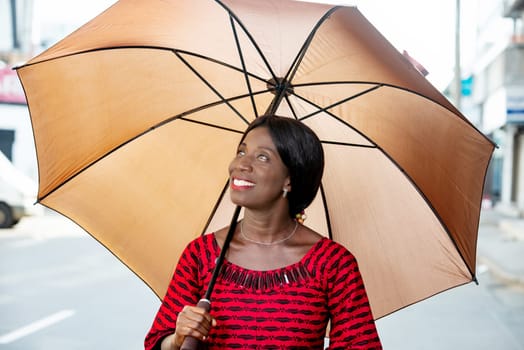 beautiful african adult businesswoman dressed in red loincloth holding umbrella in hand and looking up smiling in the street