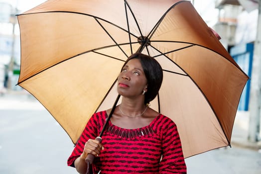 beautiful african adult businesswoman dressed in red loincloth holding umbrella in hand and looking up smiling in the street
