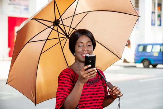 beautiful african adult businesswoman standing, holding an umbrella in hand, taking a self portrait photo in the street or chatting on video on her mobile phone smiling