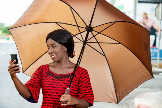 beautiful african adult businesswoman standing, holding in hand an umbrella taking a self portrait photo in the street or chatting on video on his mobile phone