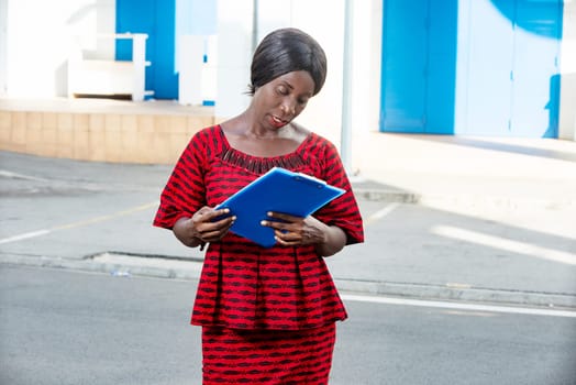 beautiful african adult businesswoman dressed in red loincloth holding folders and documents in her hands standing at modern business center in the street