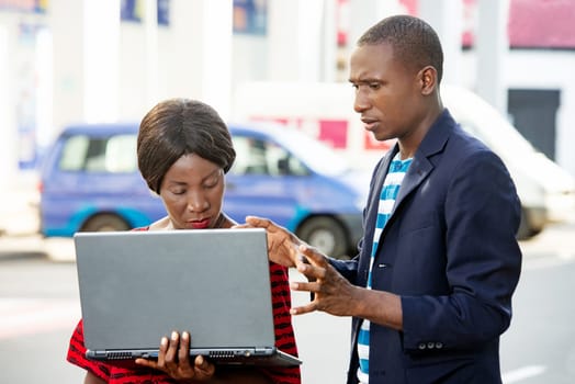 beautiful woman and handsome african adult businessman using a laptop standing, talking and displaying documents in the street