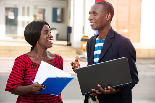 adult woman and a man working together on a project using a laptop in the street and a folders while smiling in the street