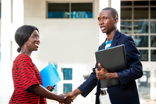 portrait of handsome businessman holding a computer and beautiful african adult business woman holding folders, shaking hands on the street smiling