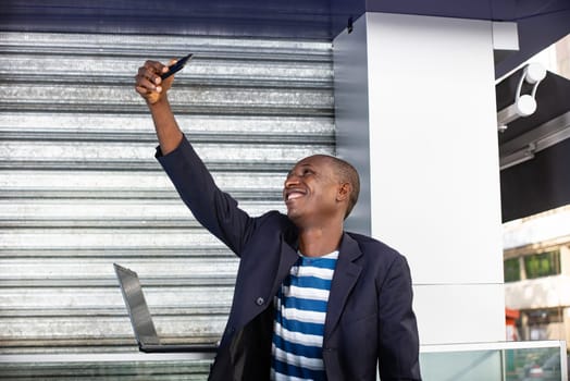 Handsome african adult man making selfie photo on cellphone camera while standing in city street in sunny summer day.