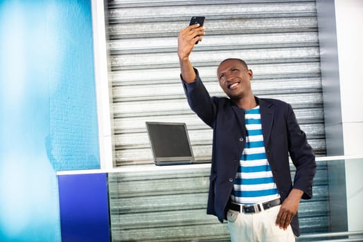 Handsome african adult man making selfie photo on cellphone camera while standing in city street in sunny summer day. Smiling man having an online video call on smartphone during free time outdoors