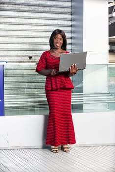 beautiful adult businesswoman dressed in red loincloth suit is standing using a laptop while being smiling