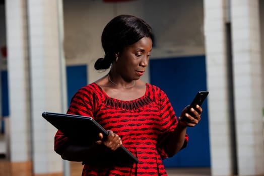 beautiful african businesswoman dressed in red loincloth standing in the street is looking at her phone holding a laptop in hand