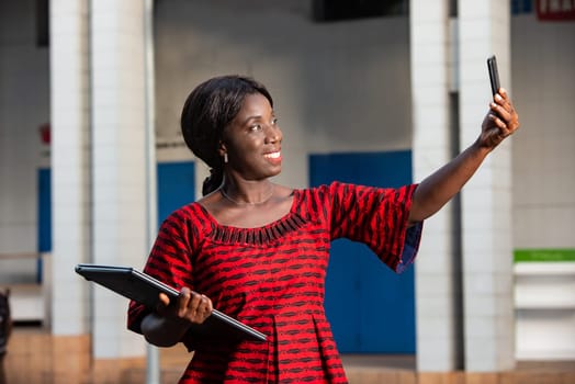 african businesswoman standing on the street is making a video call or photo in her phone holding a laptop in her hand