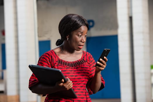 beautiful african businesswoman dressed in red loincloth standing in the street is looking at her phone holding a laptop in hand