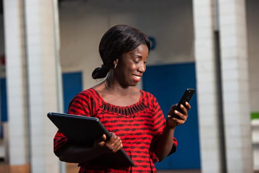 beautiful african businesswoman dressed in red loincloth standing in the street is looking at her phone holding a laptop in hand