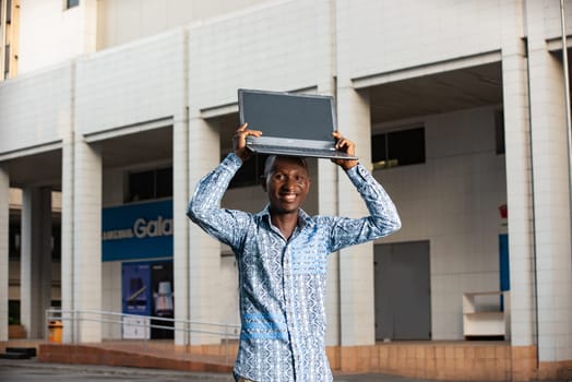 Happy african adult man standing while holding a laptop on his head in the street