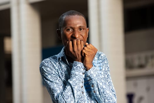 African stressed and nervous adult man with hands on mouth while biting his nails in the street