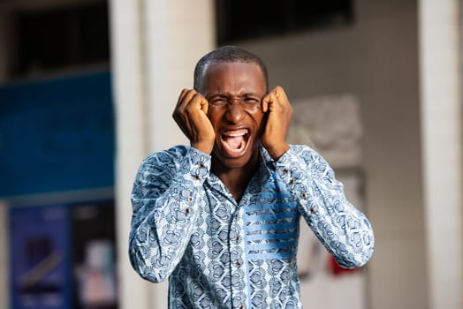 African adult man wearing a shirt is screaming with an aggressive expression covering his ears and shaking in the open air