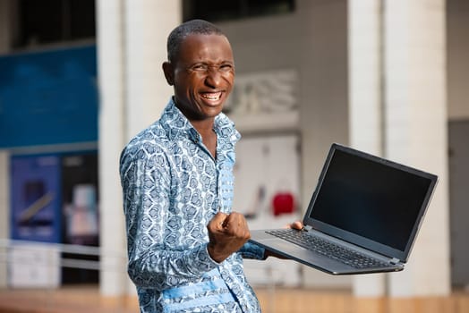 handsome happy african adult man wearing a blue shirt using a computer making a winner gesture in on the street
