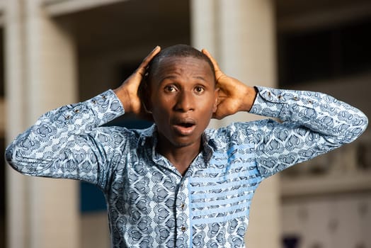 young african businessman surprised in shirt and jeans holding hands on his head in the street