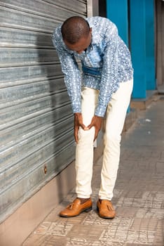 young african businessman holding knee with his hands in pain after suffering a muscle injury while walking down the street