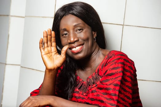 beautiful African woman in traditional dress sitting near a wall smiling at the camera while making hand gesture.