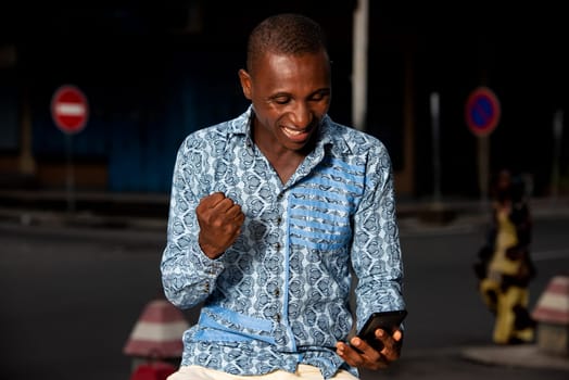 Happy african adult entrepreneur sitting in the street receiving good news in his mobile phone and celebrating his success making the winner's gesture and smiling