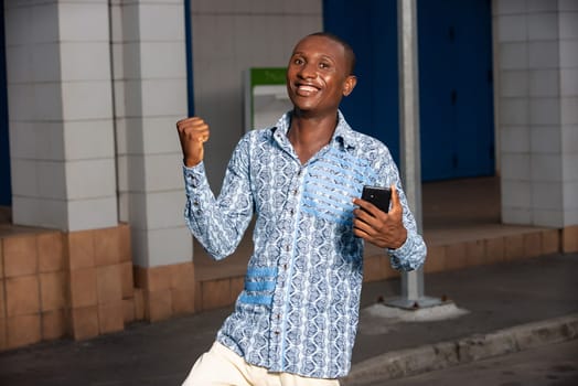 handsome african adult man standing in the street making a victory gesture with his mobile phone in hand