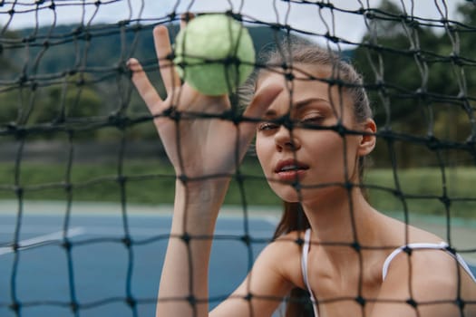 A young female tennis player captures a green tennis ball while standing behind a net, expressing focus and determination in an outdoor court environment with natural greenery