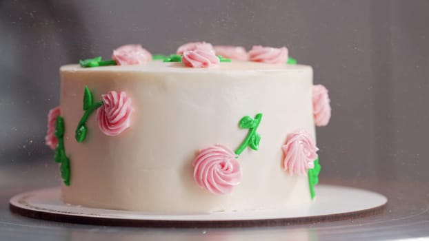 Beautiful buttercream cake on turntable. Decorated with delicate pink flowers, green leaves on smooth white icing. Bakery, celebration, wedding, pastry concept. High quality